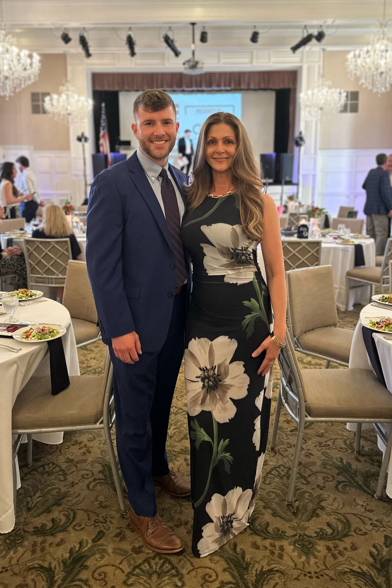 A man in a blue suit and a woman in a floral dress at a formal event with tables and chandeliers.