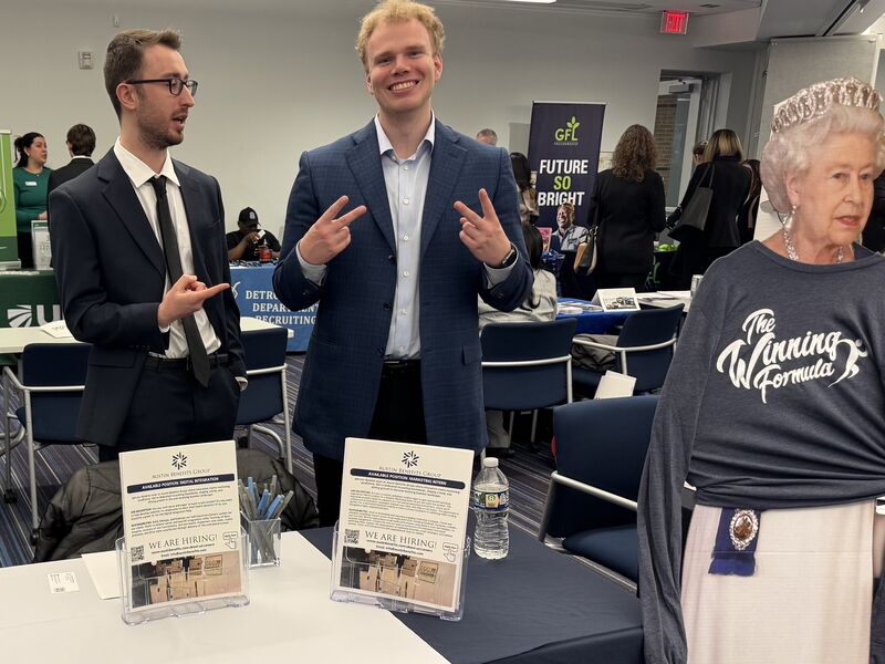 Two men in business attire smiling at a job fair booth with a cardboard cutout figure.